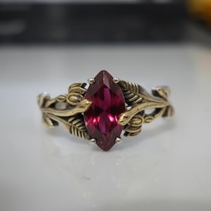 Könnte beinhalten: Ring mit einem Rubin im Marquiseschliff und einem Gold-Silber-Band. Der Ring hat einen zentralen, leuchtend roten Edelstein, der in einem detaillierten Goldfiligran-Design mit Blatt- und Blumenmotiven gefasst ist. Das Band ist aus Silber.