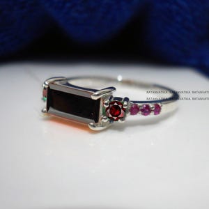 May include: A silver ring featuring a black rectangular gemstone and a red gemstone surrounded by smaller pink gemstones. The ring is displayed on a white surface.  Ratanvatika Ratanvatika Ratanvatika Ratanvatika Ratanvatika Ratanvatika