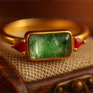 Puede incluir: Un anillo de oro con una piedra verde rectangular con talla esmeralda como pieza central. Dos gemas rojas triangulares flanquean la piedra verde. El anillo está sobre una superficie de madera.
