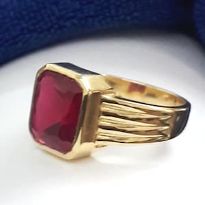 May include: A gold ring with a large, square, red gemstone. The ring has a braided band.