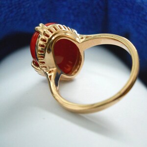May include: A gold ring with a large, oval, bright red gemstone. The ring has detailed filigree around the gemstone, and a smooth, polished gold band. The ring is set against a soft, blurred background. The ring is a piece of jewellery.