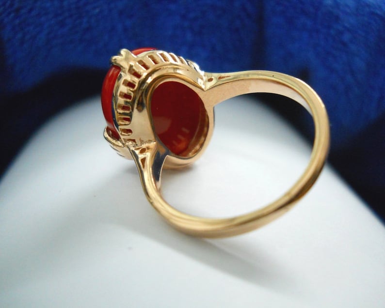May include: A gold ring with a large, oval, bright red gemstone. The ring has detailed filigree around the gemstone, and a smooth, polished gold band. The ring is set against a soft, blurred background. The ring is a piece of jewellery.
