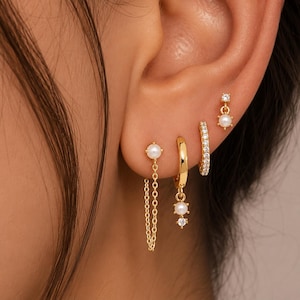 May include: A set of four gold-toned earrings worn in a single ear. The collection includes a pearl stud with a dangling chain, a small hoop with a pearl and crystal charm, a crystal-encrusted hoop, and a small stud featuring a crystal and pearl.