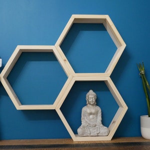 Peut inclure: Trois étagères hexagonales en bois montées sur un mur bleu. Les étagères sont empilées en quinconce. Une petite statue de Bouddha en argent est placée sur l'étagère du milieu. Une plante en pot se trouve à droite des étagères.
