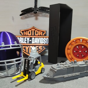 Könnte beinhalten: Eine Sammlung von 3D-gedruckten Objekten, darunter ein lilafarbener Footballhelm, ein Harley-Davidson-Logo, ein schwarzes rechteckiges Objekt, ein orangefarbenes und rotes Kugellager, ein grauer Spielzeug-Gabelstapler und ein graues Spielzeug-Kreuzfahrtschiff.