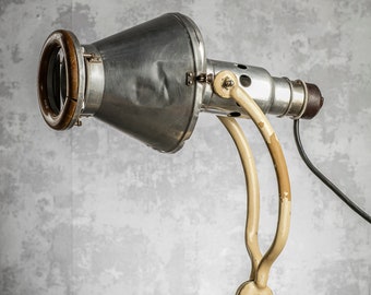 Könnte beinhalten: Eine vintage Schreibtischlampe aus Metall mit einem weißen Sockel und einem verstellbaren silbernen Arm. Die Lampe hat einen großen, runden, silbernen Reflektor mit einem Holzring um die Öffnung.