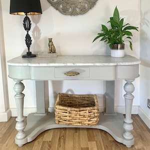 May include: A grey console table with a white marble top and a single drawer. The table has turned legs and a lower shelf. A woven basket sits on the shelf. A gold leaf mirror with a carved wooden frame hangs on the wall above the table.