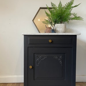 May include: A black painted wooden cabinet with a single drawer and a door. The cabinet has a carved design on the door and gold-coloured knobs. The cabinet is sitting on a wood floor.