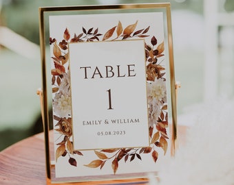 May include: A gold-framed table number card with a watercolor design of fall leaves and flowers. The card reads "TABLE 1" and "EMILY & WILLIAM 05.08.2023".