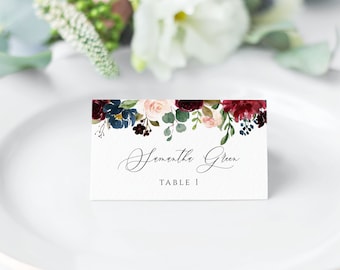 May include: A white place card with a floral design in shades of burgundy, pink, and blue. The text "Samantha Green" and "Table 1" is printed in black.