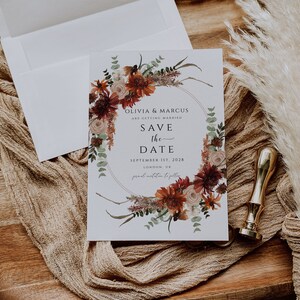 Peut inclure: Carte de sauvegarde de la date avec une couronne de fleurs dans des tons de rouge, orange et blanc. Le texte indique "Olivia & Marcus se marient. Sauvegarde de la date. 1er septembre 2028. Londres, Royaume-Uni. Invitation formelle &agrave; suivre."