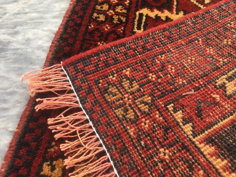 Puede incluir: Una alfombra roja, negra y beige con un borde de flecos. La alfombra tiene un dise&ntilde;o geom&eacute;trico y est&aacute; hecha de lana.