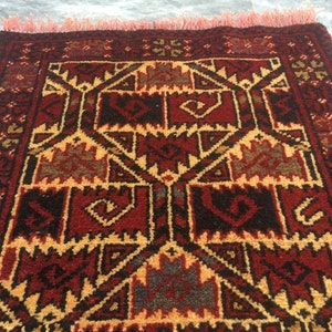 Puede incluir: Una alfombra roja, negra y dorada con un dise&ntilde;o geom&eacute;trico. La alfombra tiene un borde de flecos.