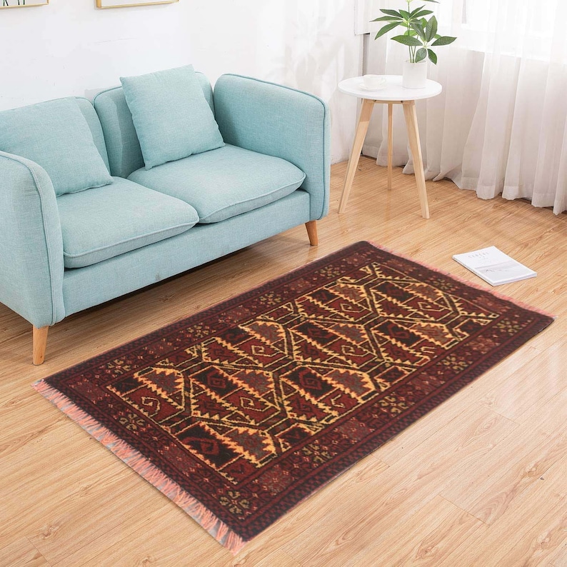 Puede incluir: Una alfombra roja, marr&oacute;n y amarilla con un patr&oacute;n y un borde de flecos. La alfombra est&aacute; colocada sobre un suelo de madera delante de un sof&aacute; azul claro.