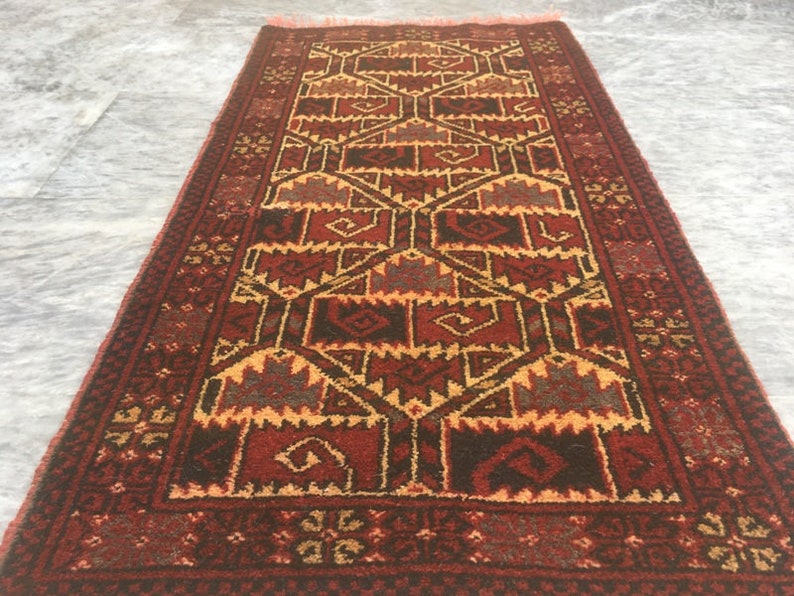 Puede incluir: Una alfombra roja, marr&oacute;n y amarilla con un dise&ntilde;o geom&eacute;trico. La alfombra tiene un borde con flecos.