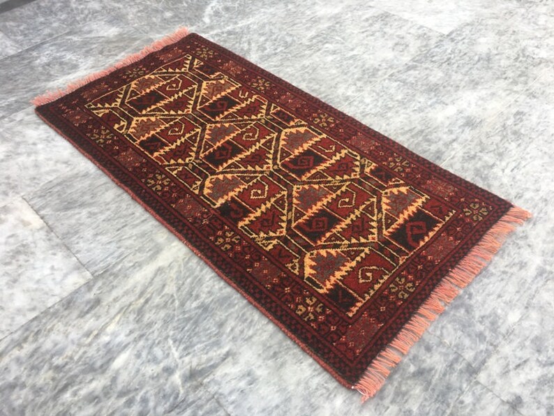 Puede incluir: Una alfombra roja, naranja y negra con un borde de flecos. La alfombra tiene un dise&ntilde;o geom&eacute;trico con un patr&oacute;n de diamantes.
