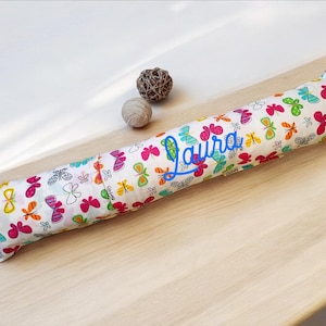 May include: A white fabric bolster pillow with a colorful butterfly pattern and the name "Laura" embroidered in blue.