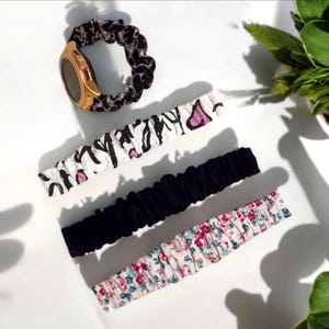 Pode incluir: Um smartwatch dourado com uma pulseira scrunchie com estampa de leopardo está acima de três pulseiras scrunchie. Uma é preta, outra tem um padrão floral e a outra tem uma estampa de zebra em preto e branco com detalhes em rosa. Em fundo branco.