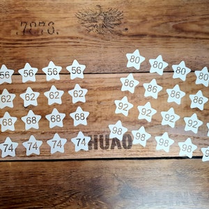 Pode incluir: Recortes de papel em forma de estrela brancos com n&uacute;meros impressos neles, variando de 56 a 98, dispostos em filas sobre uma superf&iacute;cie de madeira.