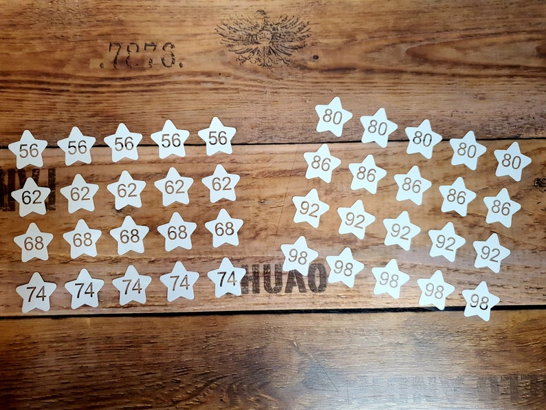 Pode incluir: Recortes de papel em forma de estrela brancos com n&uacute;meros impressos neles, variando de 56 a 98, dispostos em filas sobre uma superf&iacute;cie de madeira.