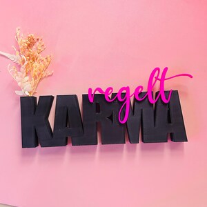 Puede incluir: Un letrero de madera negro con la palabra "KARMA" y una superposición rosa intenso "regelt", sobre un fondo rosa. Flores secas se colocan a la izquierda del letrero.