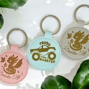 May include: Three round leather keyrings with different designs. The first keyring is light blue with a monster truck and the text "Crush Type 1 Diabetes". The second keyring is pink with a dragon and the text "Diabetes Typ 1". The third keyring is grey with a dragon and the text "Diabetes Typ 1".