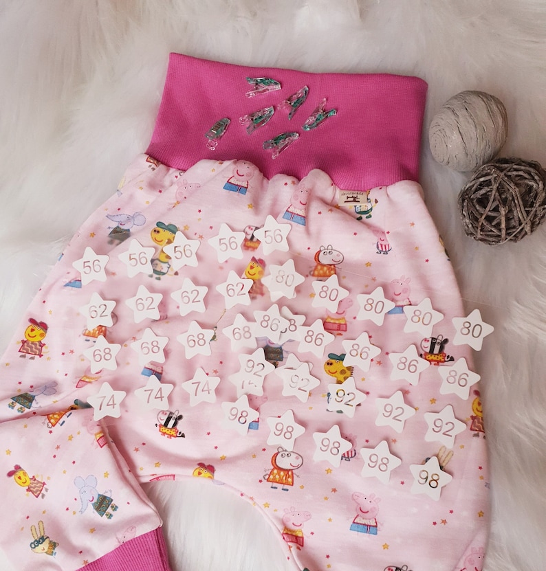 Pode incluir: Cal&ccedil;as de tecido rosa com uma cintura branca e patches em forma de estrela branca com n&uacute;meros neles. Os n&uacute;meros nos patches s&atilde;o 56, 62, 68, 74, 80, 86, 92 e 98.