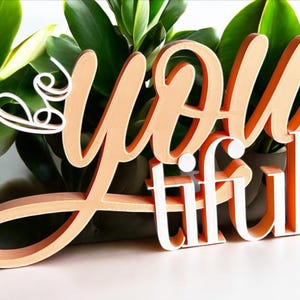 Könnte beinhalten: Ein Holzschild mit dem Schriftzug "Be You tiful" in Schreibschrift. Die Buchstaben sind pfirsichfarben mit weißen Akzenten, vor grünem Laub. Das Schild ist ein dekorativer Artikel.