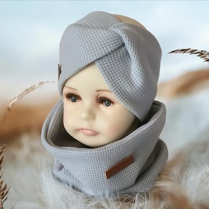 May include: A light gray knitted headband and neck warmer set. The headband has a twisted design and the neck warmer is a simple tube shape.