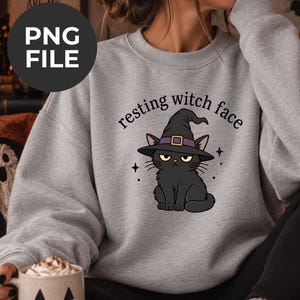 May include: A light gray crewneck sweatshirt featuring a graphic of a black cat wearing a purple-banded black witch hat. Above the cat, the text reads "resting witch face" in a black serif font, accented by small black four-pointed stars.