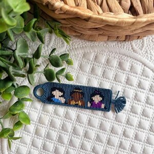 May include: A blue friendship bracelet with three small, pixelated figures in white, brown, and purple. The bracelet has a loop on one end and a tassel on the other. The background includes a woven basket and greenery.
