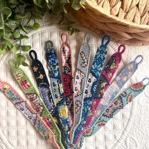 May include: A collection of handmade friendship bracelets in various colors and patterns. Each bracelet features intricate woven designs with a loop at one end. The bracelets are arranged on a white quilted surface, with a woven basket and greenery in the background.
