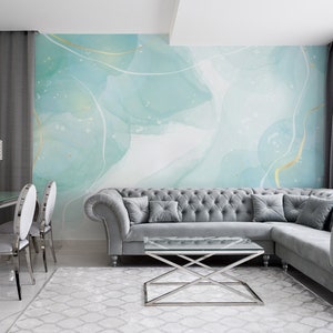 Può includere: Un interno di soggiorno con un divano angolare grigio, un tavolino da caffè in vetro e un murale astratto blu chiaro e oro. Il murale presenta linee bianche e dorate e un sottile effetto shimmer. La stanza è decorata con tende grigie e un tavolo da pranzo in vetro con sedie argentate.