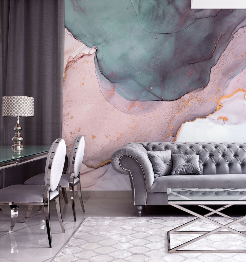 May include: A living room interior featuring a grey tufted sofa, two grey chairs, a glass-topped coffee table, and a lamp with a patterned shade. The wall behind the furniture is adorned with a large abstract painting in shades of pink, green, and gold.