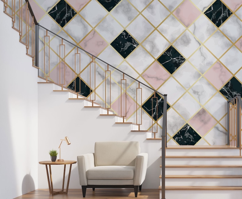 May include: A modern interior featuring a staircase with a geometric marble wall design. The wall has a pattern of white, pink, and black marble squares outlined with gold lines. A cream-colored armchair and small wooden table with a lamp and plant are in the foreground.