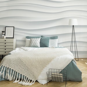 Puede incluir: Una cama blanca con una manta de punto blanco y una falda de cama azul turquesa. La cama tiene almohadas blancas con un patrón geométrico y almohadas azul turquesa. Hay un revistero de metal negro en el suelo al lado de la cama. La pared detrás de la cama tiene un patrón de ondas blancas en 3D.