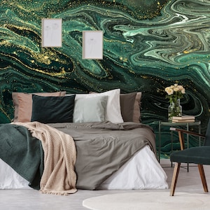 Op de afbeelding: Een slaapkamer met een groene en gouden abstracte muurschildering. Het bed heeft een wit dekbed, een groene deken en verschillende kussens. Een donkergroene fauteuil staat in de hoek van de kamer.