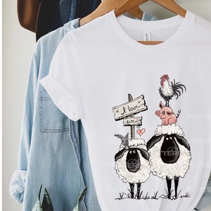 以下が含まれることがあります： 白いTシャツに、2匹の羊、豚、鶏のグラフィックが描かれています。羊は緑の芝生の上に立っています。豚は鶏の背中に乗っています。鶏は「愛してる」と書かれた木製の看板の上に立っています。