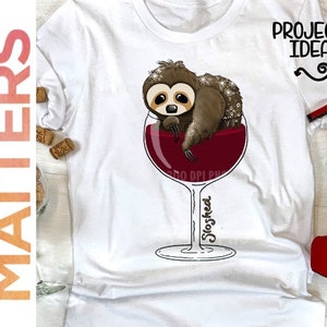 May include: White t-shirt featuring a graphic of a sloth in a glass of red wine. The text "Sloshed" is written on the glass. The shirt is lying on a wooden surface with a pair of red high heels.