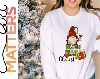 Op de afbeelding: Wit sweatshirt met een kerstmannetje ontwerp. De kerstmannetje zit op een stapel cadeaus en houdt een glas wijn vast. De tekst "Cheers!" is onder de kerstmannetje gedrukt.