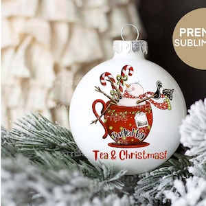 K&ouml;nnte beinhalten: Wei&szlig;er Weihnachtsbaumschmuck mit einem Schneemann in einer roten Teetasse. Der Schneemann tr&auml;gt einen roten Schal und h&auml;lt einen Zuckerstock. Der Text auf dem Ornament lautet "Fueled by Tea & Christmas!"