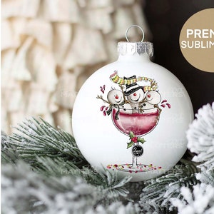 May include: White Christmas ornament with three snowmen in a wine glass. The snowmen are wearing hats and scarves. The wine glass is filled with red wine and has a holly sprig on the stem. The ornament is surrounded by a snowy pine branch.