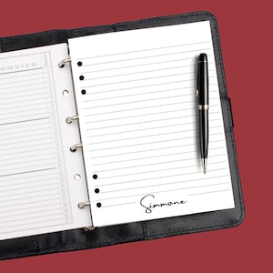 May include: A black leather bound planner with a white page open. The page has lines and a black pen is lying on the page. The name "Simmone" is written in black ink at the bottom of the page.