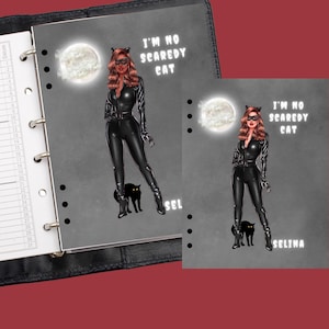 May include: A gray planner with a black leather cover and a black and white illustration of a woman in a black catsuit with a black cat. The text "I'm no scaredy cat" is written in white on the planner. The planner has a ring binder and a date column.