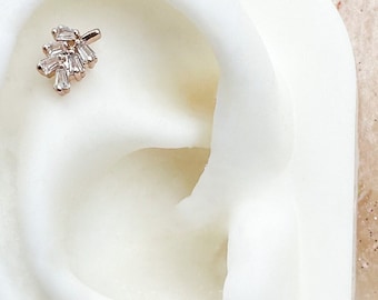 Puede incluir: Primer plano de un modelo de oreja blanco con un pequeño pendiente de oro en forma de diamante con un grupo de diamantes en forma de baguette.