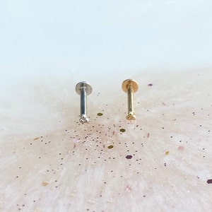 Puede incluir: Dos anillos de nariz con un diseño de tres bolas. El anillo de la izquierda es plateado y el anillo de la derecha es dorado.