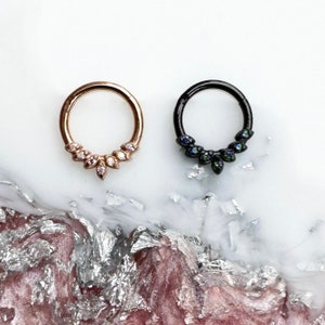 May include: Two hoop earrings. One is rose gold with a cluster of small, clear gemstones. The other is black with a cluster of iridescent gemstones. Both earrings are on a white and pink surface.
