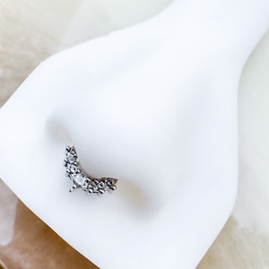 May include: A silver-toned nose ring with a curved design. The jewelry features a series of small, clear gemstones and a larger, central stone. The nose ring is displayed on a white surface.