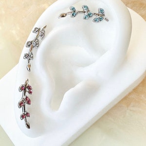 May include: Three silver ear cuffs with crystal accents. The ear cuffs are shaped like a branch with leaves and feature pink, blue, and clear crystals.