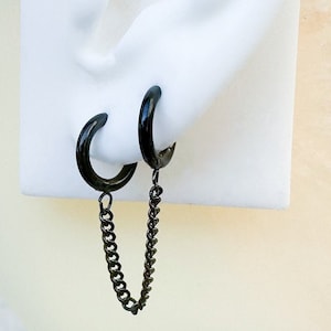 Puede incluir: Un par de pendientes de aro negros conectados por una cadena negra. Los pendientes se llevan en una cabeza de maniquí blanca.